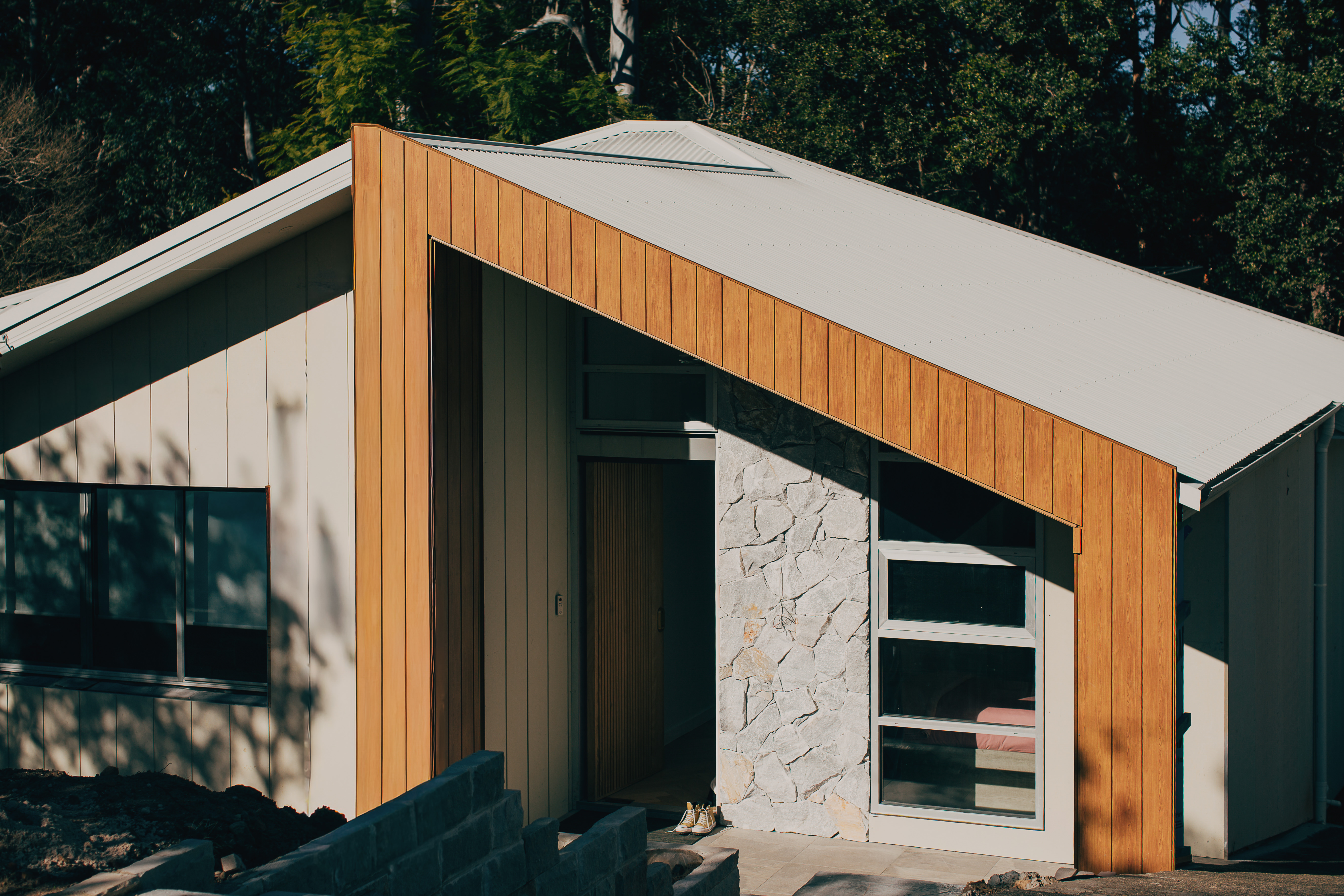 The transformed Castle Hill home exterior with an angular white roofline, timber-clad entry, a natural stone accent wall and pale weatherboard cladding against bushland